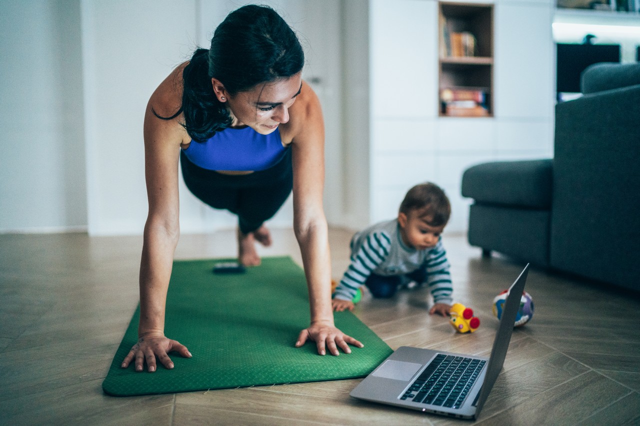 Woman exercises while watching workout video on a laptop and her baby playing around