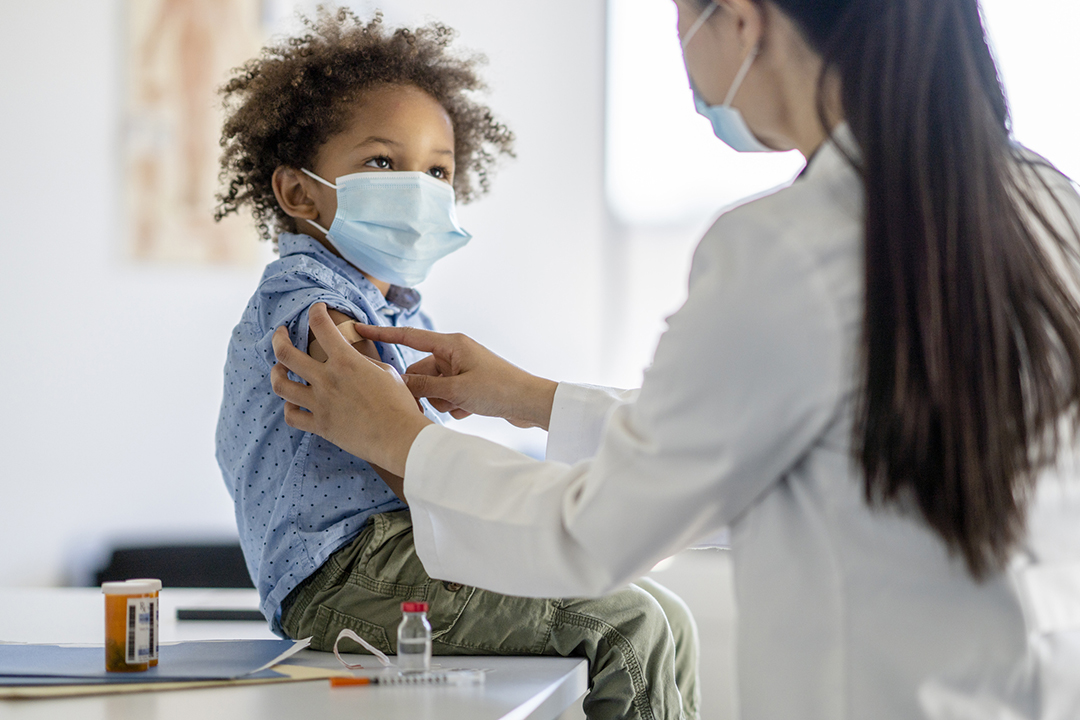 A sweet little mixed race boy sits up on an examination table as he receives an immunization. He is looking up and smiling as the doctor places the bandage on his arm. Both are wearing medical masks to protect them from COVID.