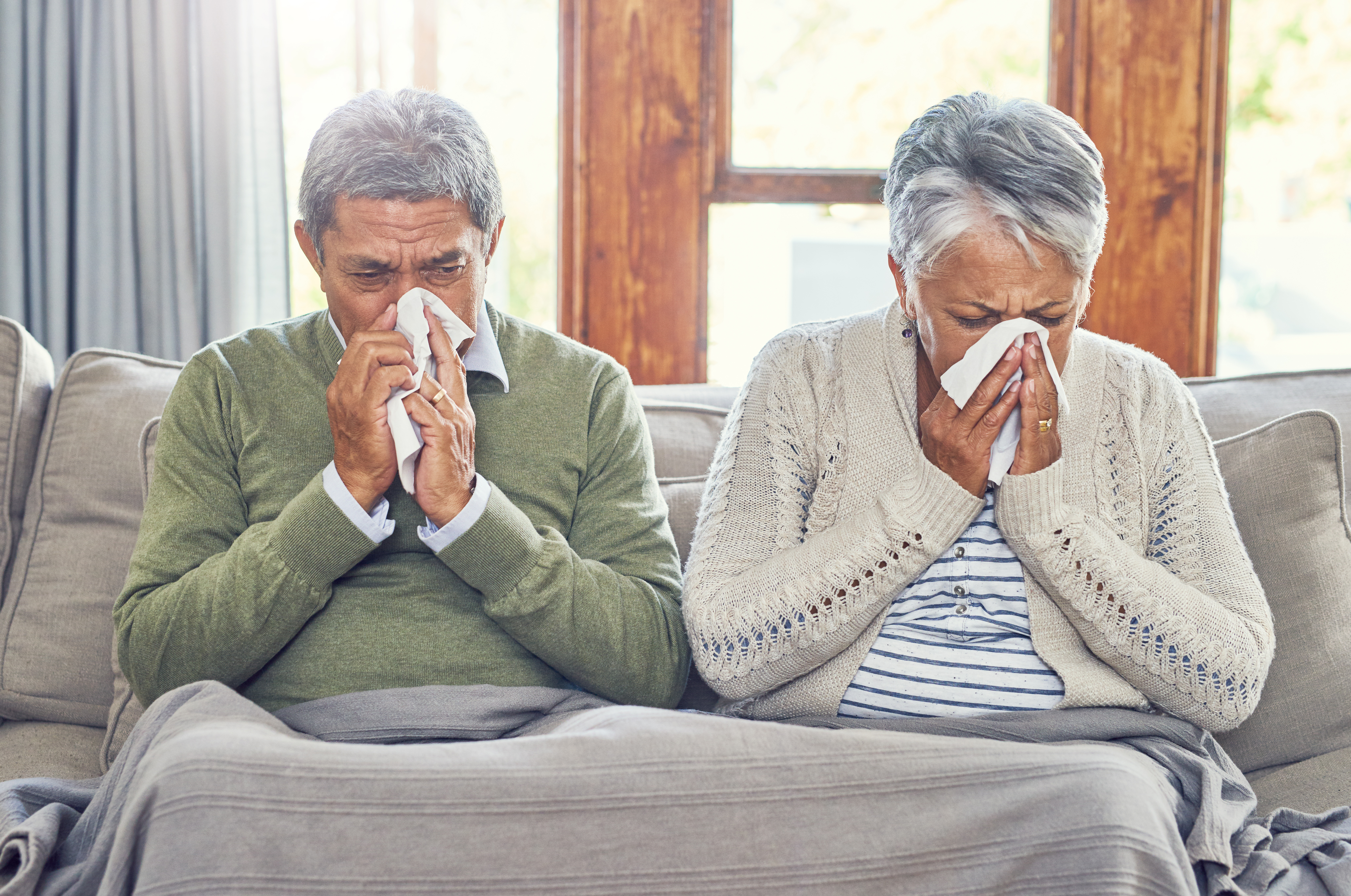 Shot of a sickly senior couple each blowing their nose with a tissue while sitting on a couch at home