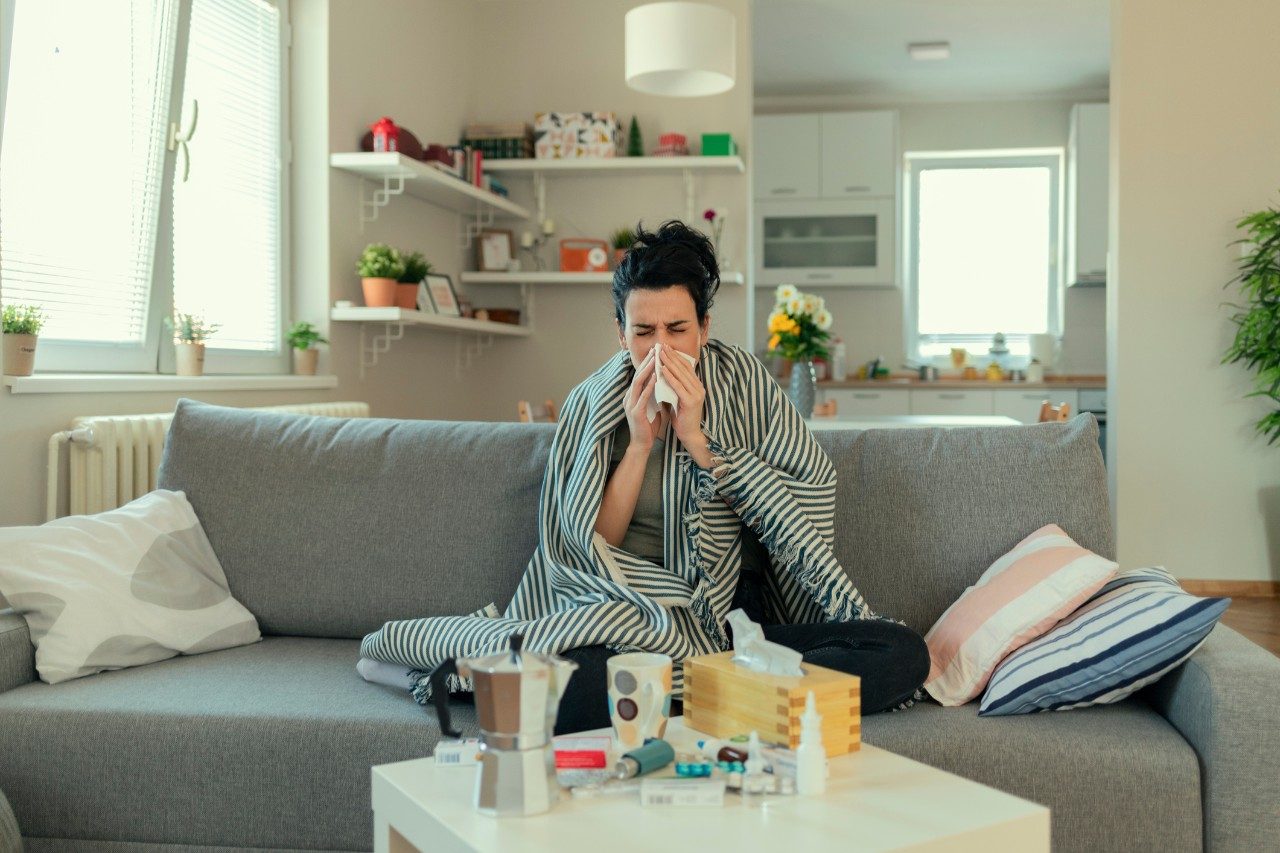 Sick Woman Covered With a Blanket Lying in Bed With High Fever and a Flu, Resting at Living Room. She Is Exhausted and Suffering From Flu. Sick Woman With Runny Nose Lying in Bed. Girl Suffering From Cold Lying in Bed With Tissue Blowing Her Nose While Sitting on the Sofa