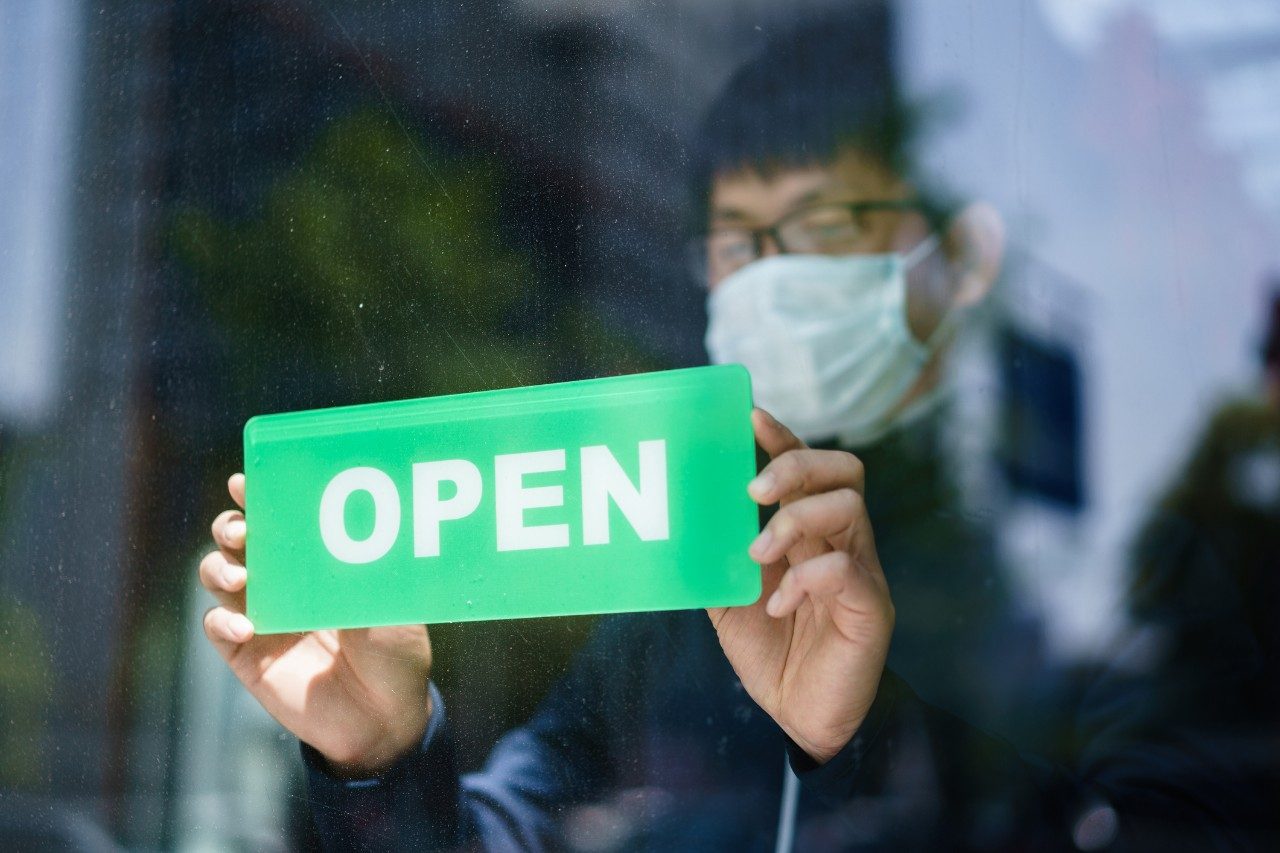 Happy business owner hanging an open sign while wearing protective mask