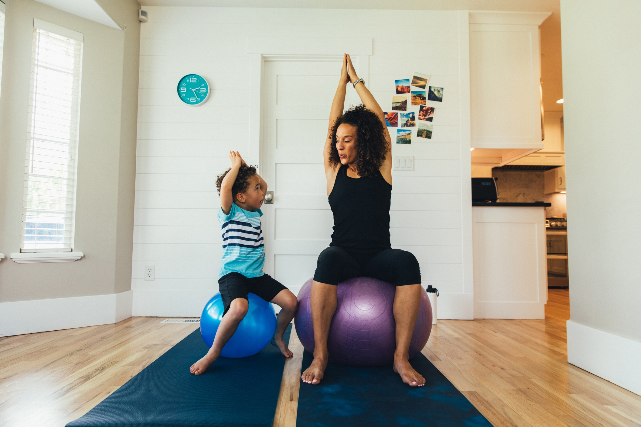 A mother exercises on a fitness ball with her young son inside their home. She is teaching the boy the importance of a healthy lifestyle by proper stretching and exercising.