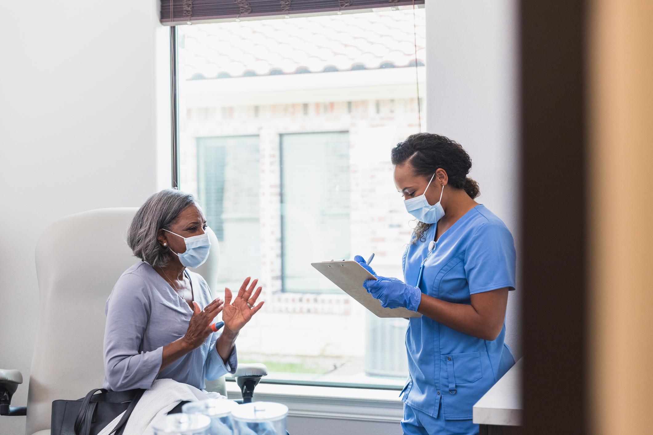 A worried senior woman gestures as she talks about troubling symptoms with a female healthcare professional. The healthcare professional takes notes as the woman talks.