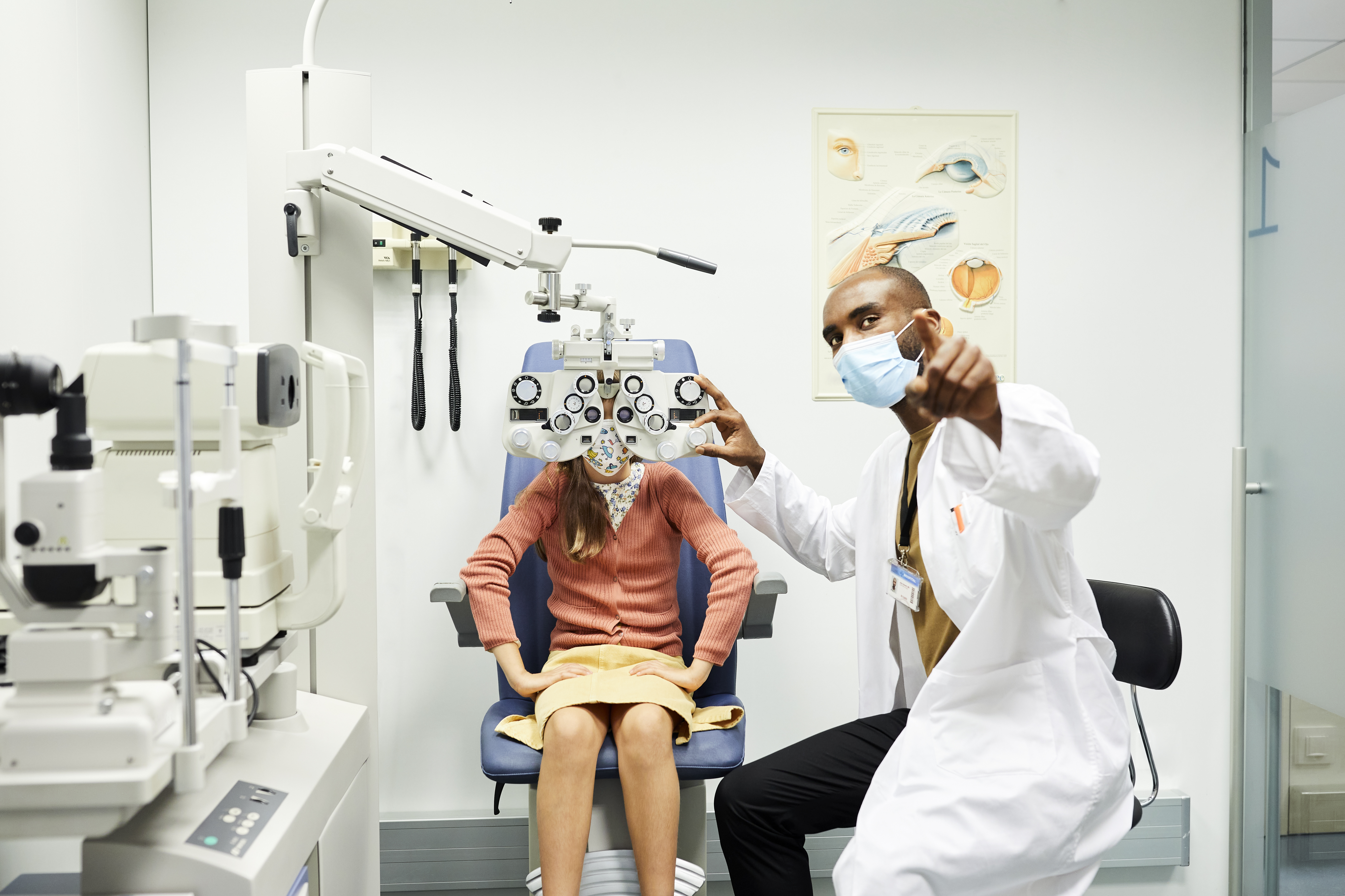 Ophthalmologist gesturing to girl while wearing protective face mask. Female child is sitting behind optometry in doctor's office. They are in hospital for eye exam during COVID-19 crisis.