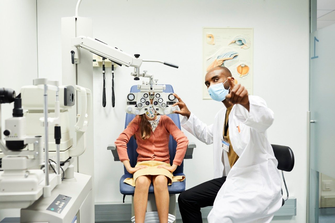 Ophthalmologist gesturing to girl while wearing protective face mask. Female child is sitting behind optometry in doctor's office. They are in hospital for eye exam during COVID-19 crisis.