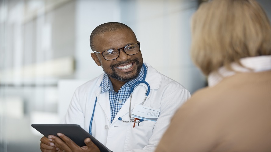 Smiling male doctor holding digital tablet while looking at senior woman. Patient is visiting healthcare worker in hospital. He is wearing lab coat and eyeglasses.