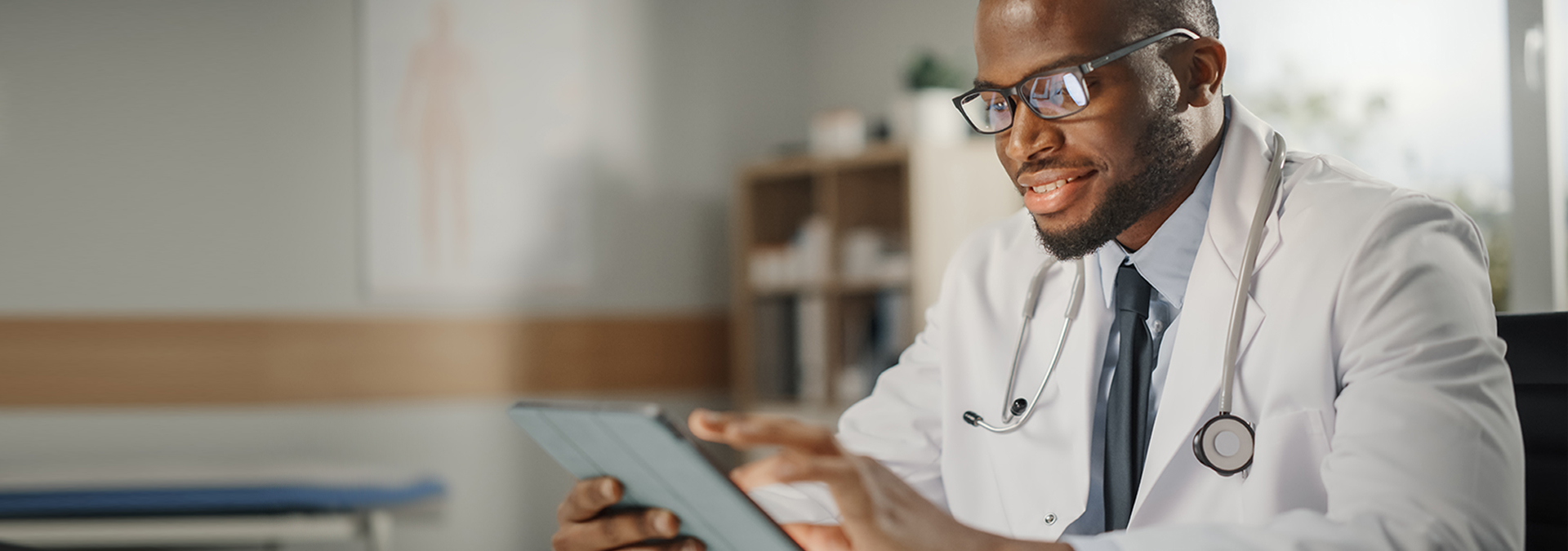 Happy and Smiling African American Male Doctor Wearing White Coat Working on Tablet Computer at His Office. Medical Health Care Professional Working with Test Results, Patient Treatment Planning.