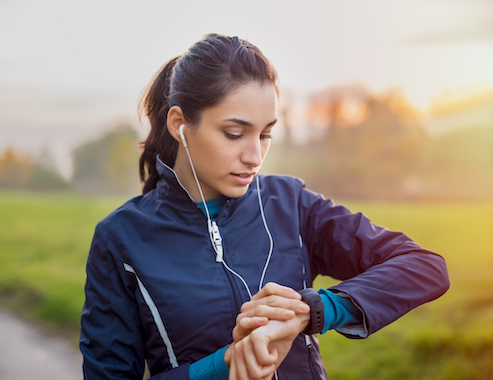 Young athlete listening to music during workout at park and adjusting smart watch. Young latin woman setting smartwatch before jogging in winter or autumn.