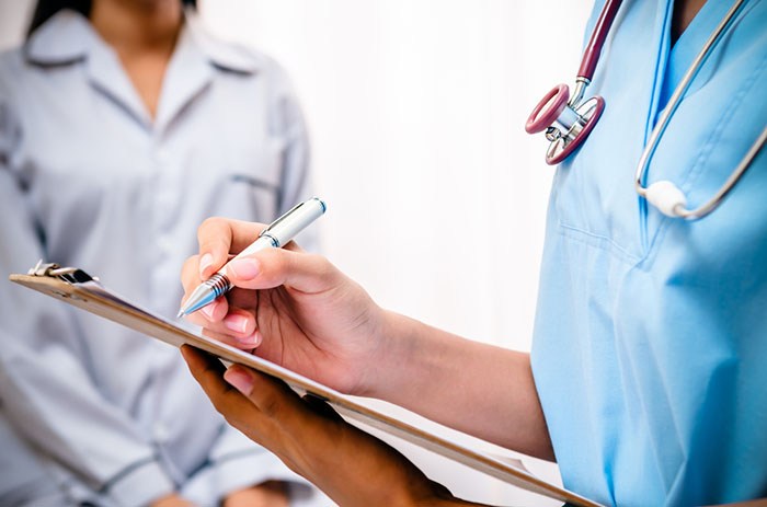 Female doctor taking notes on clipboard with female patient in background.