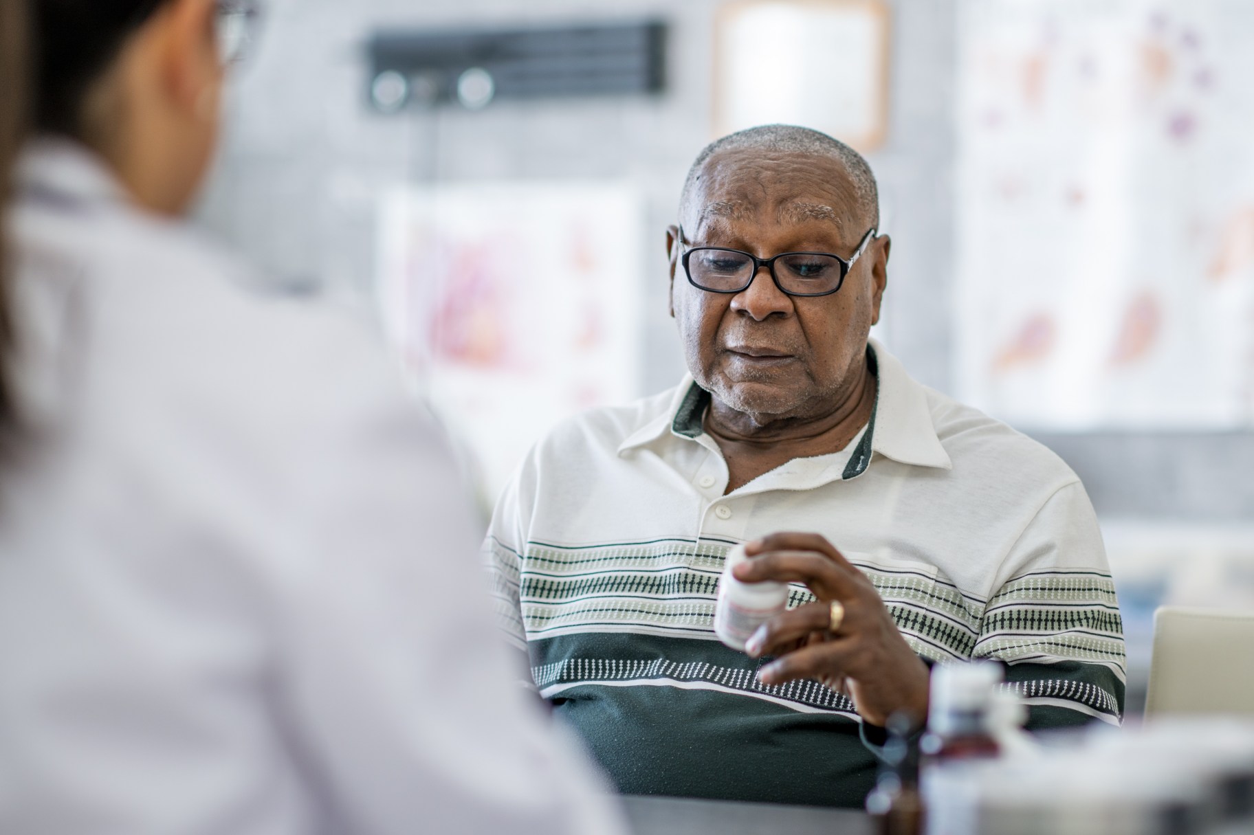 An elderly man looks at a pill bottle in his hand while sitting across the table from a doctor in the doctor's office. He appears to be making a decision about it.