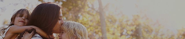 Woman giving child piggyback ride while kissing other child on forehead.