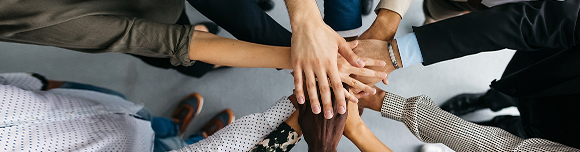 A high-angle shot of a group of male and female colleagues putting their hands together in an office. They are dressed in fashionable business clothes. Their faces are not visible, only their arms. Horizontal daylight indoor photo.