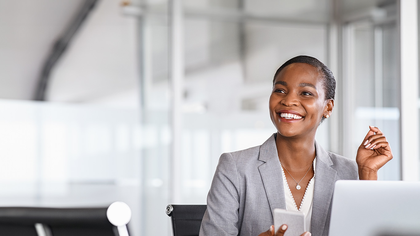 African black business woman using smartphone while working on laptop at office. Smiling mature african american businesswoman lookingup at copy space while working on phone. Successful woman entrepreneur think about new business ideas.