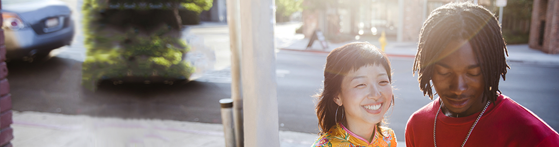 a young black man and Asian woman standing on a street sidewalk talking and smiling back lit