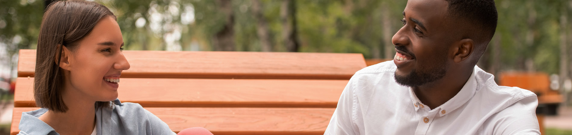 Happy young intercultural couple in casualwear sitting on wooden bench in park or square, chatting and enjoying summer day at leisure