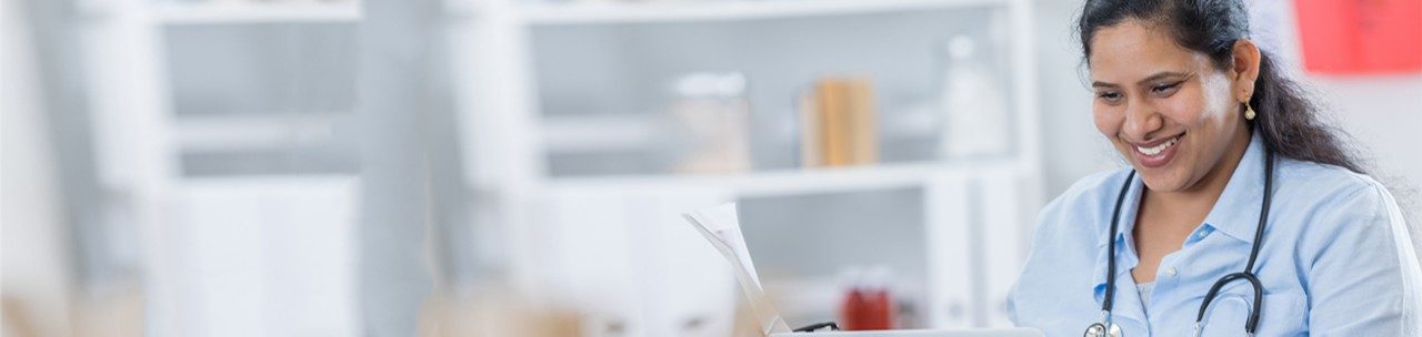 Pretty mid adult Indian doctor uses a laptop to research something while working in her office. She is smiling confidently while using the computer. She is wearing a stethoscope.